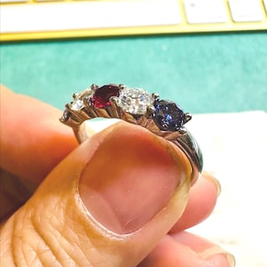 May include: A silver ring featuring a series of gemstones. The ring showcases a sequence of stones: a small white stone, a ruby, a large clear stone, and a sapphire. The ring is held by a person's hand.