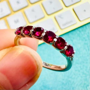 May include: A silver ring with six vibrant red gemstones. The gemstones are oval-shaped and set closely together, creating a continuous line of color. The ring is held up against a light blue background.
