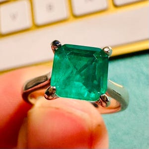 May include: A square-cut emerald ring with a silver band. The vibrant green gemstone is held in place by four prongs. The ring is set against a blurred background of a keyboard and a light blue surface.