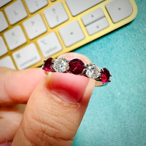 Peut inclure: Une bague en argent ornée d'alternances de rubis ronds et de diamants. Les pierres précieuses sont serties de près, créant une bande continue de couleur et d'éclat. La bague est tenue sur un fond flou.