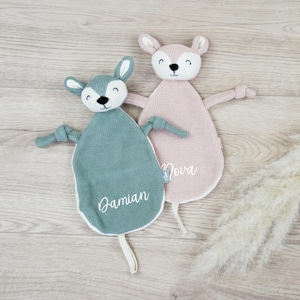 Könnte beinhalten: Zwei gestrickte Baby-Schmusetücher in Form eines Rehs. Eines ist türkis mit dem Namen "Damian" in Weiß gestickt, das andere rosa mit "Nova". Beide haben weiße Akzente. Ideal für Neugeborene.