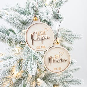 Könnte beinhalten: Zwei hölzerne Weihnachtskugeln hängen an einem schneebedeckten Baum. Eine Kugel trägt die Aufschrift "Bester Papa" mit "Deine Mara" darunter, die andere "Beste Mama" mit "Dein Theo". Jede hat eine kleine goldene Glocke.