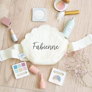 Könnte beinhalten: Eine Sammlung von Holzspielzeug-Schminkartikeln, darunter eine muschelförmige Tasche mit dem Namen "Fabienne", Lippenstifte, Lidschattenpaletten, ein Kompaktspiegel, Rouge und eine Fixierspray-Flasche. Auf einer hellen Holzoberfläche angeordnet.