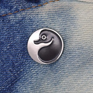 May include: A black and white yin yang pin with a stylized bird design. The pin is on a blue denim background.
