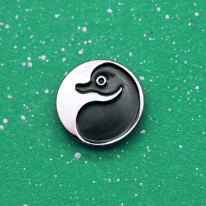 May include: A silver pin with a black and white yin yang design. The yin side features a stylized duck head.