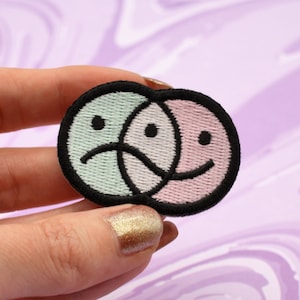 May include: An embroidered patch featuring two overlapping faces. One face is green and has a sad expression, the other face is pink and has a happy expression.