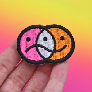 May include: Embroidered patch with a split design of two smiley faces, one sad and pink, the other happy and orange, with black outlines. The patch is held against a vibrant, colourful gradient background.