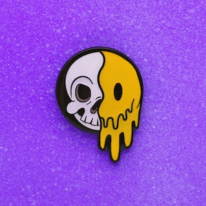 May include: A round enamel pin featuring a split design: a white skull on the left and a melting yellow smiley face on the right. The pin has a black border and is set against a purple background.