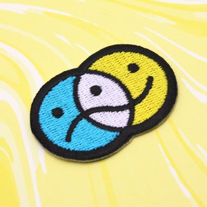 May include: A blue and yellow embroidered patch with two faces, one smiling and one frowning, representing duality.