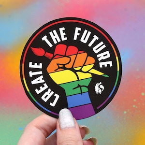 May include: A round sticker with a black background and white text that reads "Create the Future". A rainbow fist with a paintbrush is in the center of the sticker.