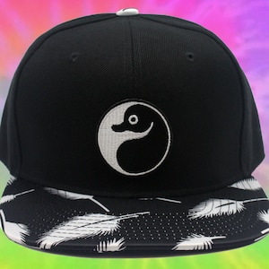 May include: Black and white baseball cap with a yin yang symbol featuring a duck. The brim of the cap has a black and white feather pattern.
