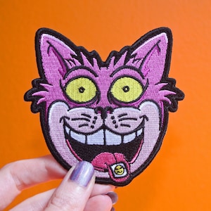 K&ouml;nnte beinhalten: Gestickter Aufn&auml;her mit einem grinsenden Katzengesicht, mit gro&szlig;en gelben Augen und einer rosa Zunge mit einem Smiley. Die Katze ist rosa mit schwarzer Umrandung. Der Aufn&auml;her befindet sich auf einem orangefarbenen Hintergrund.