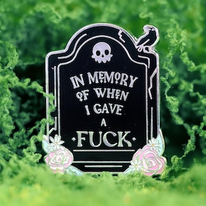May include: Black enamel pin in the shape of a tombstone with the text "IN MEMORY OF WHEN I GAVE A FUCK". The pin features a skull, a bird, and floral accents in pink and white. The pin is set against a green background.