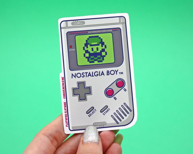 May include: A sticker of a gray and white Game Boy with a green screen displaying a pixelated character. The sticker has the text "NOSTALGIA BOY TM" on it.