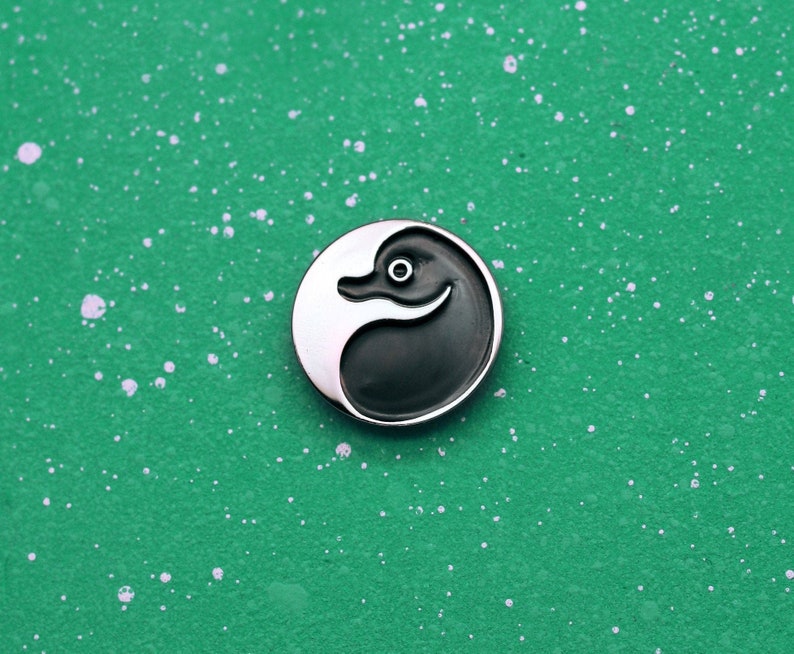 May include: A silver pin with a black and white yin yang design. The yin side features a stylized duck head.