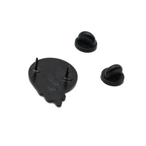 May include: Black enamel pin with two pin backs. The pin is a stylized shape with two posts for attachment. The pin backs are black, with a butterfly clutch style.