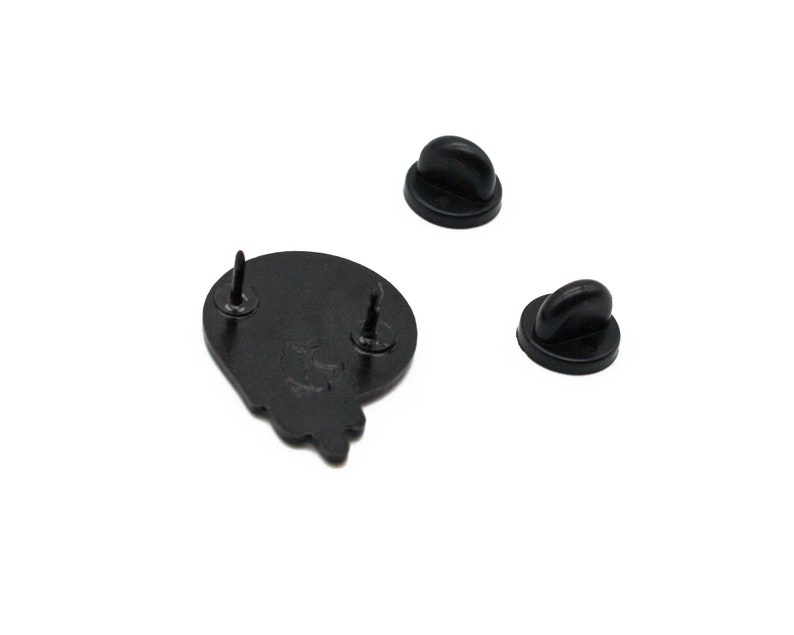 May include: Black enamel pin with two pin backs. The pin is a stylized shape with two posts for attachment. The pin backs are black, with a butterfly clutch style.