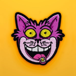 K&ouml;nnte beinhalten: Gestickter Aufn&auml;her mit einem Cartoon-Katzenkopf mit breitem Grinsen, gelben Augen und einer rosa Zunge mit Smiley. Die Katze ist lila mit schwarzer Umrandung, vor einem leuchtend gelben Hintergrund.