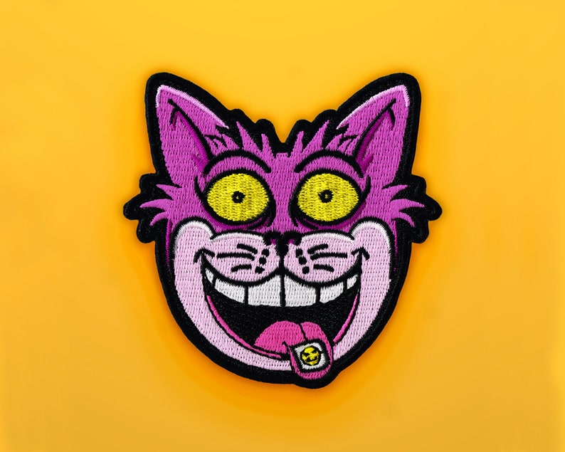 K&ouml;nnte beinhalten: Gestickter Aufn&auml;her mit einem Cartoon-Katzenkopf mit breitem Grinsen, gelben Augen und einer rosa Zunge mit Smiley. Die Katze ist lila mit schwarzer Umrandung, vor einem leuchtend gelben Hintergrund.