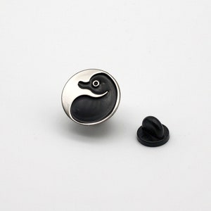 May include: A silver and black enamel pin featuring a duck design. The pin has a black rubber clutch back.