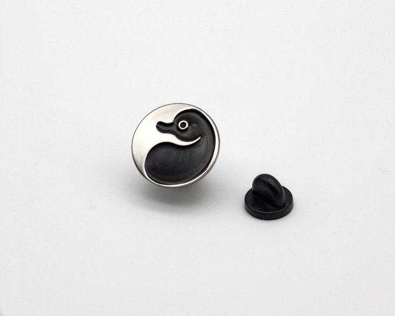 May include: A silver and black enamel pin featuring a duck design. The pin has a black rubber clutch back.