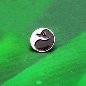 May include: A black and white enamel pin featuring a stylized duck in a yin yang design.