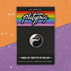 May include: A black and white enamel pin featuring a cartoon platypus with a black body and white head. The pin is on a black and white background with the text "Platypus" in white and the text "Soft Enamel Pin" and "Made by Misfits in the UK" in gray. The text "@platypus_official | www.platypus.clothing" is also included in gray.