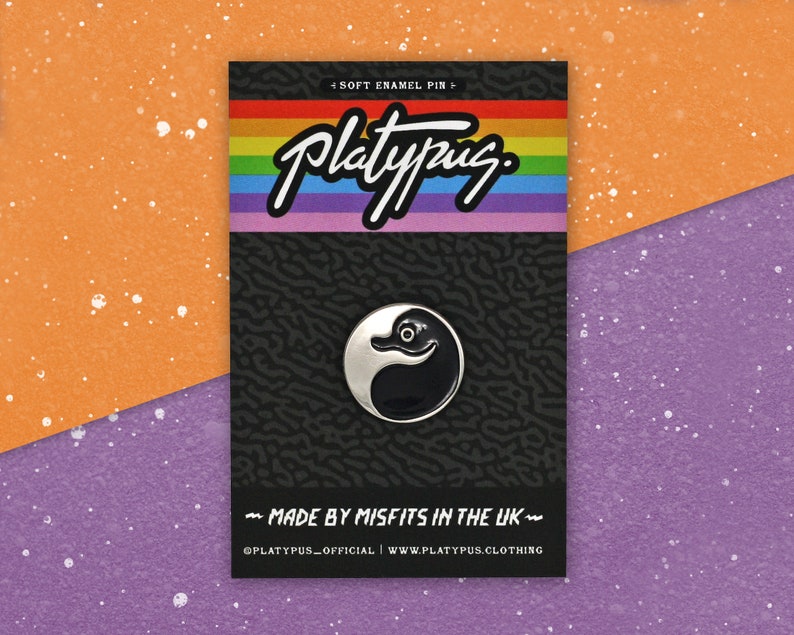 May include: A black and white enamel pin featuring a cartoon platypus with a black body and white head. The pin is on a black and white background with the text "Platypus" in white and the text "Soft Enamel Pin" and "Made by Misfits in the UK" in gray. The text "@platypus_official | www.platypus.clothing" is also included in gray.