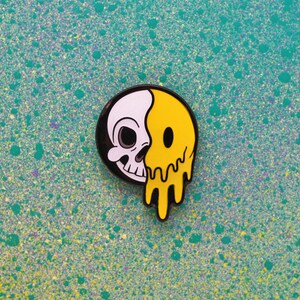 May include: A round enamel pin featuring a split design: one half is a white skull, the other half is a yellow smiley face with a dripping effect. The pin has a black border and is set against a teal background.