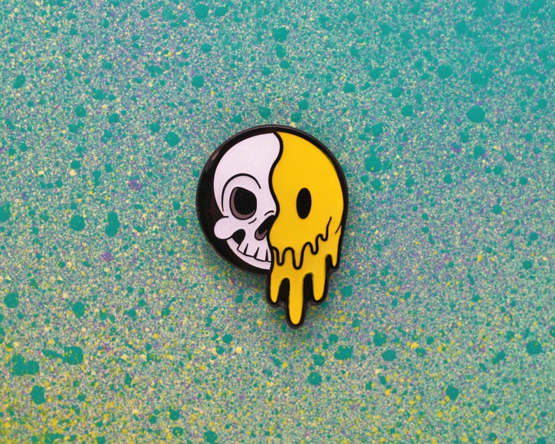May include: A round enamel pin featuring a split design: one half is a white skull, the other half is a yellow smiley face with a dripping effect. The pin has a black border and is set against a teal background.