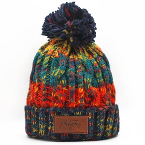 Puede incluir: Un gorro de punto colorido con un pompón en la parte superior. El gorro tiene un parche de cuero marrón con el texto "Platypus" en él.