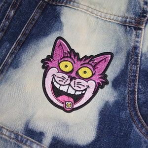 K&ouml;nnte beinhalten: Gestickter Aufn&auml;her mit einem grinsenden Cartoon-Katzenkopf mit rosa Fell, gelben Augen und schwarzem Umriss. Die Zunge der Katze hat ein kleines, rundes, gelb-schwarzes Design. Der Aufn&auml;her ist auf einer Jeansjacke befestigt.