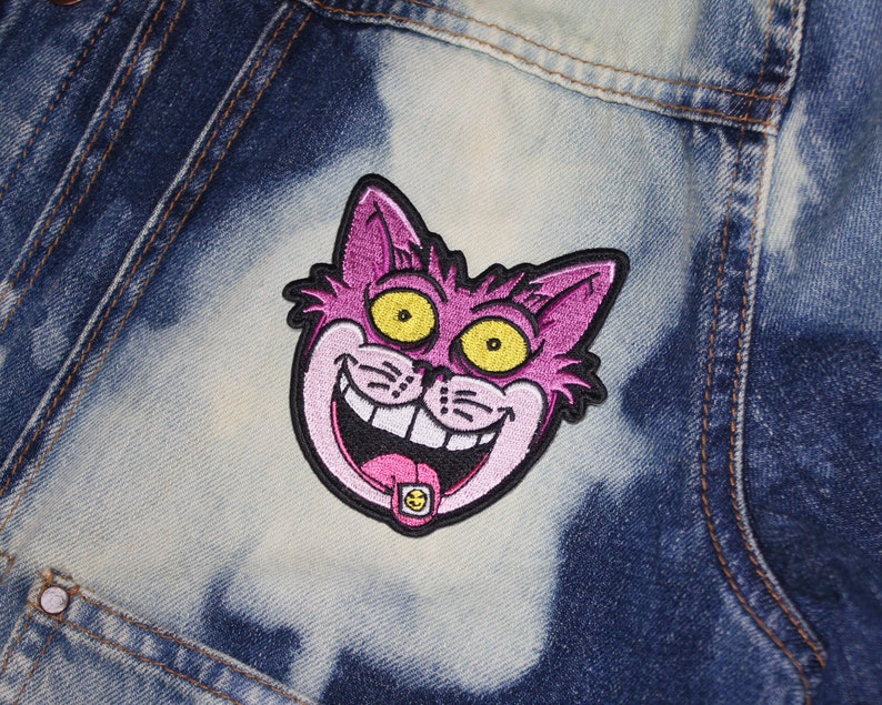 K&ouml;nnte beinhalten: Gestickter Aufn&auml;her mit einem grinsenden Cartoon-Katzenkopf mit rosa Fell, gelben Augen und schwarzem Umriss. Die Zunge der Katze hat ein kleines, rundes, gelb-schwarzes Design. Der Aufn&auml;her ist auf einer Jeansjacke befestigt.