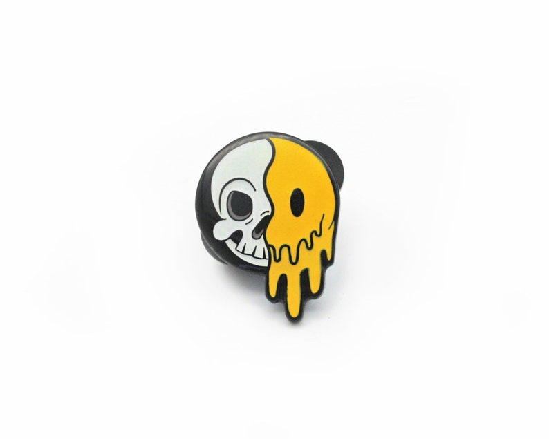 May include: A black and white skull and yellow smiley face enamel pin. The pin features a half skull and half melting smiley face design. The pin is a fun accessory for jackets, bags, or hats.