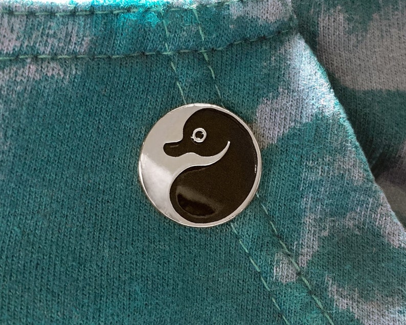 May include: A silver pin with a black and white yin yang design featuring a stylized duck head.