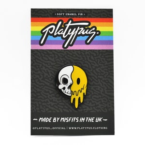 May include: A soft enamel pin featuring a half-skull, half-melting yellow smiley face design. The pin is on a black backing card with the word "Platypus" in white script above a rainbow stripe. Text on the card reads "MADE BY MISFITS IN THE UK".