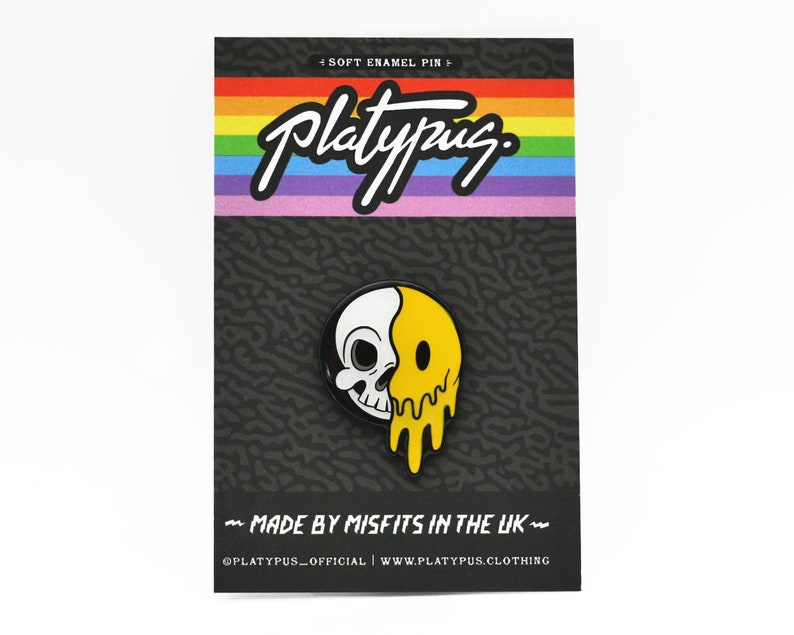 May include: A soft enamel pin featuring a half-skull, half-melting yellow smiley face design. The pin is on a black backing card with the word "Platypus" in white script above a rainbow stripe. Text on the card reads "MADE BY MISFITS IN THE UK".