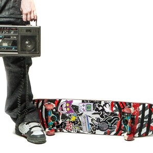 May include: A person wearing jeans and sneakers holds a vintage cassette player with a long cord. A skateboard with red, black, and white stripes and various stickers is on the floor.