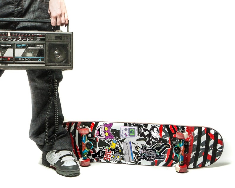 May include: A person wearing jeans and sneakers holds a vintage cassette player with a long cord. A skateboard with red, black, and white stripes and various stickers is on the floor.