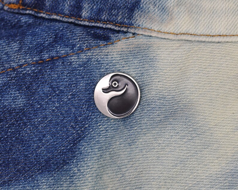 May include: A black and white yin yang pin with a stylized bird design. The pin is on a blue denim background.
