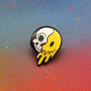 May include: A black, white, and yellow enamel pin featuring a skull design. The skull is split in half, with one side white and the other yellow with a dripping effect. The pin has a black border.