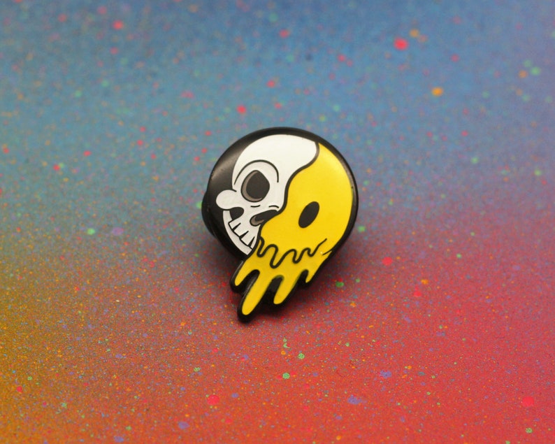 May include: A black, white, and yellow enamel pin featuring a skull design. The skull is split in half, with one side white and the other yellow with a dripping effect. The pin has a black border.