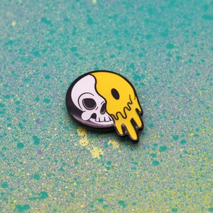 May include: A black, white, and yellow enamel pin featuring a skull design. The skull is split in half, with one side white and the other yellow and dripping. The pin is set against a teal background with paint splatters.