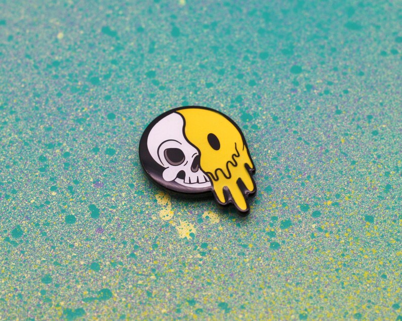 May include: A black, white, and yellow enamel pin featuring a skull design. The skull is split in half, with one side white and the other yellow and dripping. The pin is set against a teal background with paint splatters.