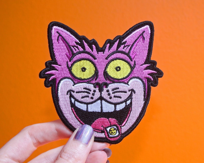 K&ouml;nnte beinhalten: Gestickter Aufn&auml;her mit einem grinsenden Katzengesicht, mit gro&szlig;en gelben Augen und einer rosa Zunge mit einem Smiley. Die Katze ist rosa mit schwarzer Umrandung. Der Aufn&auml;her befindet sich auf einem orangefarbenen Hintergrund.