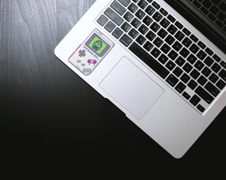May include: A silver laptop computer with a black keyboard and a sticker of a retro video game console with the text "NOSTALGIA BOY" on the top of the trackpad.