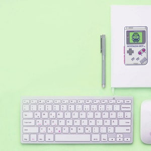 May include: A white wireless keyboard with a white mouse and a white notebook with a black and white illustration of a retro video game console with the text "NOSTALGIA BOY" on a light green surface.