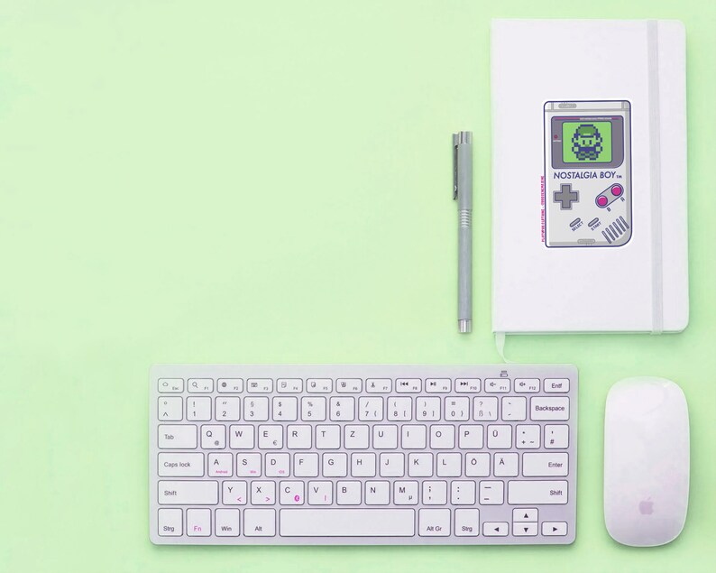 May include: A white wireless keyboard with a white mouse and a white notebook with a black and white illustration of a retro video game console with the text "NOSTALGIA BOY" on a light green surface.