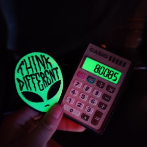 May include: A green glow-in-the-dark patch with the text "Think Different" and an alien face. A pink calculator with a green display showing the number 80085.