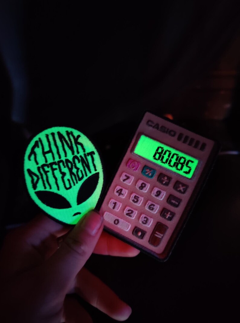 May include: A green glow-in-the-dark patch with the text "Think Different" and an alien face. A pink calculator with a green display showing the number 80085.